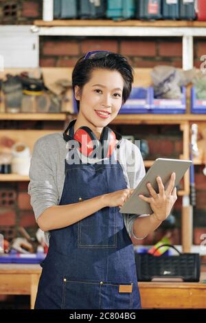 Portrait of smiling pretty young Asian carpenter in denim apron using applciation on tablet computer to check order from clients Stock Photo