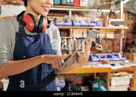 Cropped image of smiling female carpenter checking picture on tablet computer in search of inspiration for making new furniture item Stock Photo