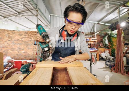Young Asian female carpenter using drill when assembling bookshelf or bookcase Stock Photo