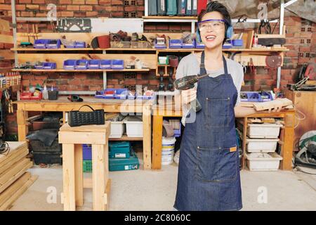 Joyful young Asian female carpenter in protective glasses posing with screwdriver in hand Stock Photo