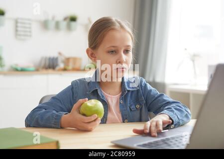 Portrait of cute girl using laptop and eating apple while studying online at home in cozy interior , copy space Stock Photo