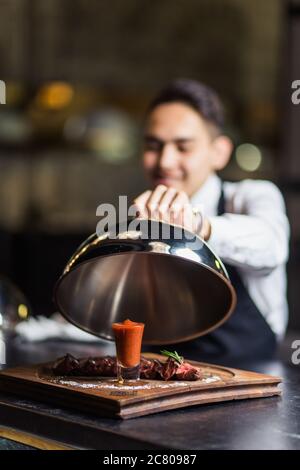 Happy young waiter in tuxedo and bowtie lifting metal cloche from serving tray Stock Photo