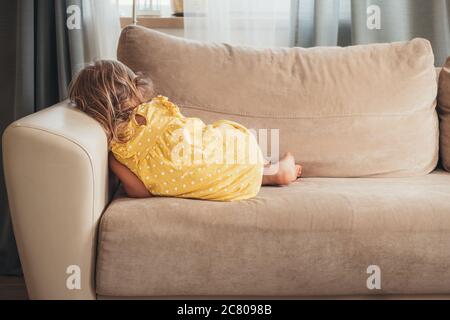 Young cute girl in a yellow dress is lying on the sofa back view, she is sad. Stock Photo