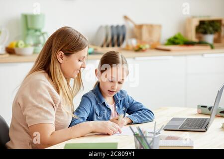 Warm-toned portrait of caring mother helping cute girl doing homework or studying at table in cozy home interior, copy space Stock Photo