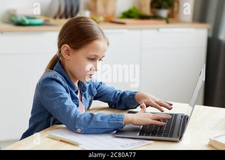 Side view portrait of cute blonde girl typing on laptop keyboard while doing homework or studying online at home, copy space Stock Photo