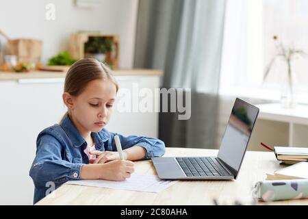 Portrait of cute blonde girl writing in notebook while doing homework or studying at home in cozy interior, copy space Stock Photo
