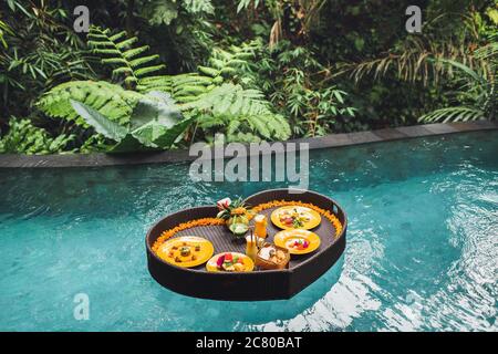 Floating breakfast in jungle swimming pool, tropical resort. Black rattan tray in heart shape, Valentines day or honeymoon surprise, view from above. Exotic summer diet. Tropical beach lifestyle. Stock Photo