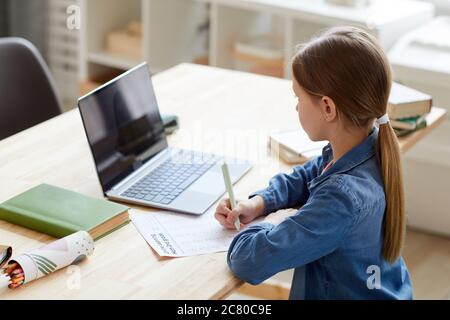High angle portrait of little girl using laptop during online class with tutor or teacher while studying at home sitting at desk in cozy interior, cop Stock Photo