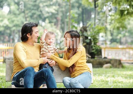 Happy young Vietnamese man and woman sitting on bench in park and adjusting sweater of thier little son Stock Photo