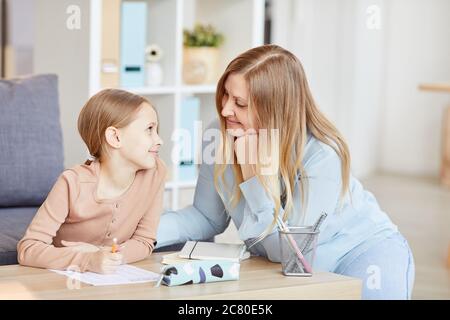Portrait of loving adult mother looking at cute little girl doing homework while studying at home in cozy interior, copy space Stock Photo