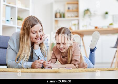 Warm-toned portrait of smiling mother and daughter lying on carpet together while drawing or studying in cozy home interior, copy space