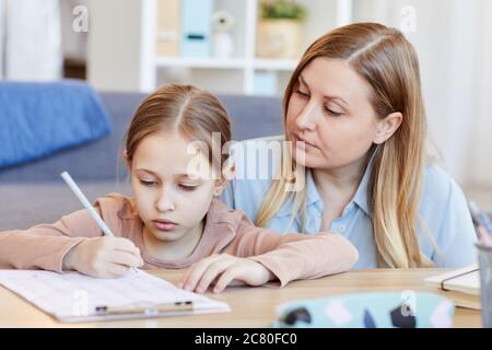 Portrait of loving adult mother looking at cute little girl doing homework or test while studying at home in cozy interior Stock Photo