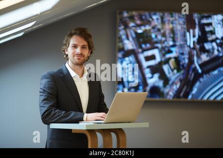 Portrait of handsome bearded businessman looking at camera and smiling while posing at standing table in futuristic office interior with city map in b Stock Photo