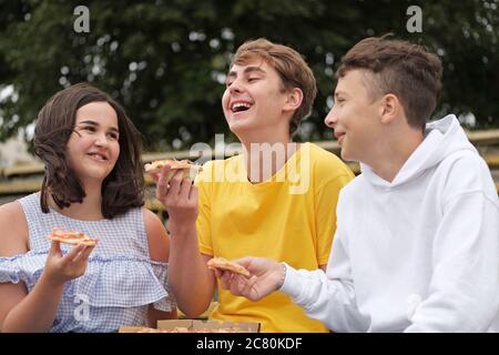 Two teenage boys and a girl sharing snacks outdoors laughing and joking together as they eat in a close up portrait Stock Photo