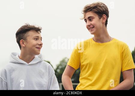 Two happy young teenage boys or brothers standing chatting outdoors in a close up low angle view against a grey high key sky Stock Photo