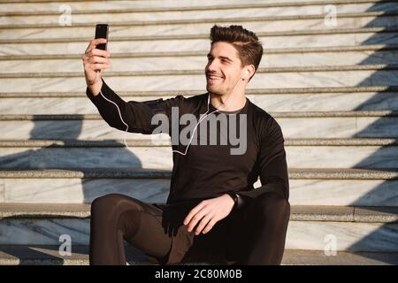 Young attractive smiling sporty man happily taking selfie on cellphone on stairs after workout outdoor Stock Photo