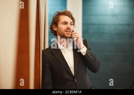 Waist up portrait of handsome bearded businessman speaking by smartphone and looking away while leaning on wall in minimal office interior, copy space Stock Photo