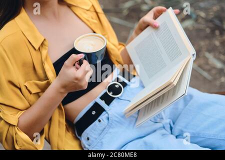 Close up woman in yellow shirt and jeans holding cup of coffee and book in hands in park Stock Photo