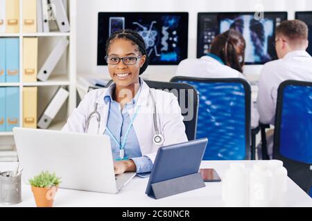 Portrait of pretty smiling young Black female doctor working on laptop at her desk Stock Photo