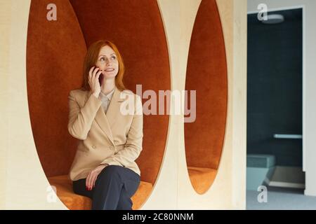 Portrait of mature red-haired businesswoman speaking by smartphone and smiling while sitting in lounge zone of modern office or coworking space Stock Photo