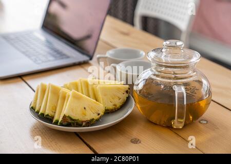 Home office workplace with open laptop, sliced pineapple and a tea mug on wooden desk Stock Photo