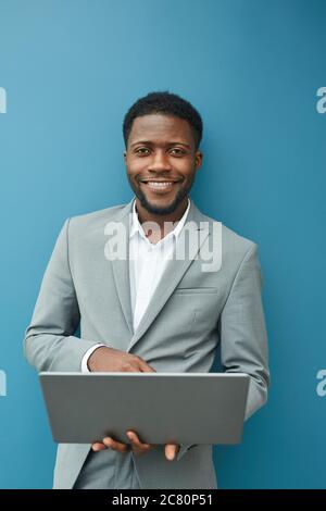 Vertical waist up portrait of contemporary African-American businessman holding laptop and smiling at camera while standing against blue wall in offic Stock Photo