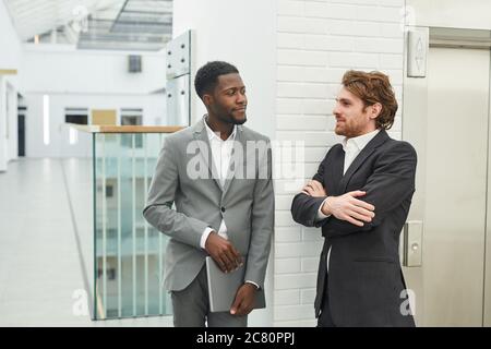 Waist up portrait of two successful businessmen wearing suits chatting while standing in while office building interior, copy space Stock Photo