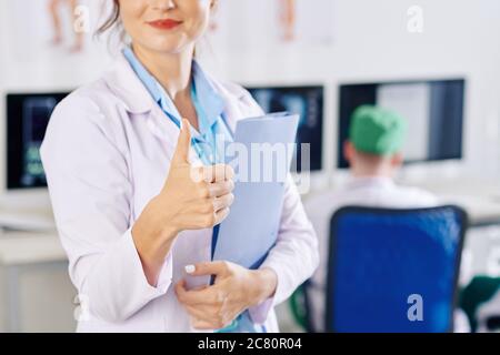 Positive young female general practitioner with documents folder in hands showing thumbs-up Stock Photo