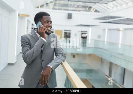 Waist up portrait of successful African-American businessman speaking by smartphone and smiling while leaning on balcony railing in modern office buil Stock Photo