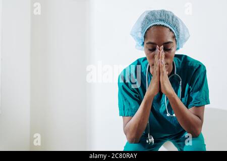Tired young Black medical nurse praying for sick and suffering people when sitting on the floor in hospital corridor Stock Photo