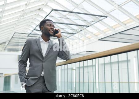Low angle portrait of confident African-American businessman speaking by smartphone and smiling while leaning on balcony railing in modern office buil Stock Photo