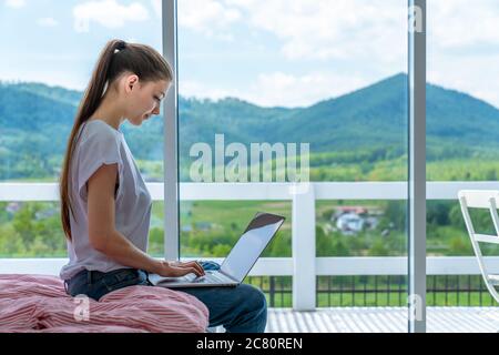 Cropped shot view of young woman keyboarding on laptop computer with blank copy space screen while sitting on bed at home, brunette female student Stock Photo