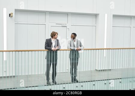 Wide angle full length view at two successful businessmen wearing suits looking at each other and smiling while standing on glass balcony in contempor Stock Photo