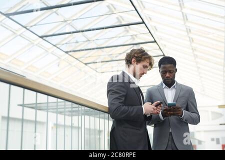 Waist up portrait of two successful businessmen wearing suits using smartphones while standing on glass balcony in contemporary office building under Stock Photo