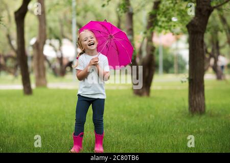 Happy laughing chil girl 2-3 year old wearing waterproof boots and holding pink umbrella posing in park over green grass at background. Happiness. Loo Stock Photo