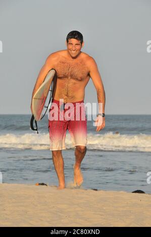 Portrait of a young handsome man in his twenties coming out of the ocean carrying a surfboard under his arm and smiling Stock Photo