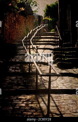 Sturdy metal railings guide visitors along a sunlit cobblestone pathway in a quaint village; France Stock Photo