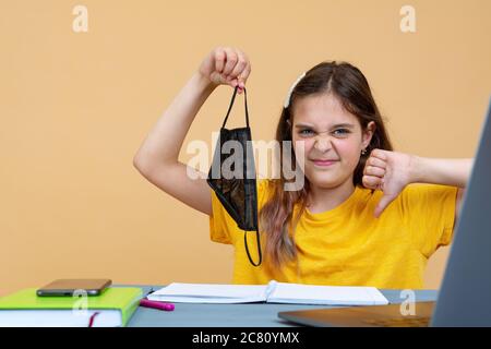 Little kid schoolgirl 12-13 years old holds face mask in her hand, study at desk with laptop isolated on orange background. School distance education Stock Photo