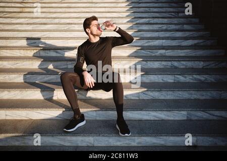Young attractive sporty man sitting on stairs while drinking water after workout outdoor Stock Photo