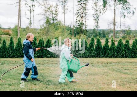 brother and sister playing in the rain with a hose and watering can Stock Photo