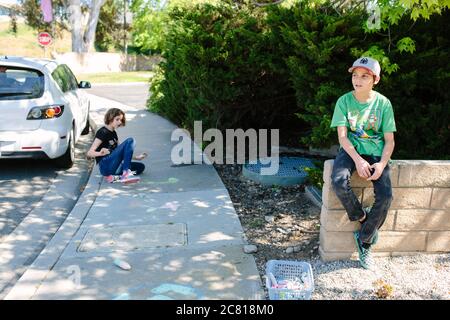 Boy sits on a wall while his sister does chalk art on the sidewalk Stock Photo