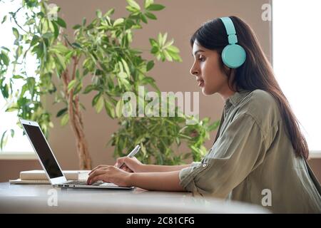 Indian teen girl student wear headphones watching online class making notes. Stock Photo