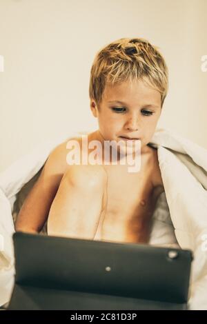 Boy with tablet watch cartoon under blanket in bed in the morning or evening night, when parents busy or sleep. Dependence on TV, difficulties to cont Stock Photo