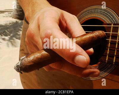 Close-up of man holding a burning cigar and playing the guitar outside, in the midday sunlight. Male street artist playing guitar. Stock Photo