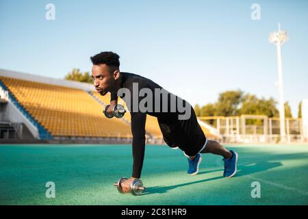 Young strong serious African American sportsman doing exercises with dumbbells at city stadium Stock Photo