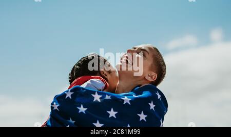 Two american female athletes wrapped in national flag giving a hug to another after winning the competition. Excited sportswomen embracing each other Stock Photo