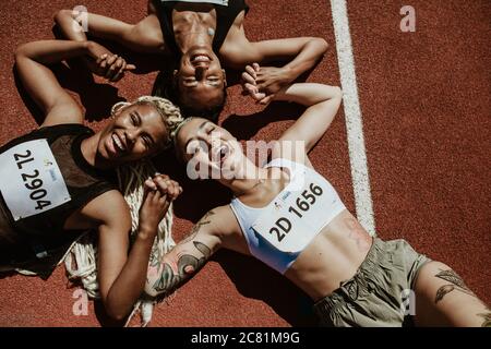 Top view of three female athletes lying together on running track holding hands and smiling while looking at camera. Successful team of runners enjoyi Stock Photo