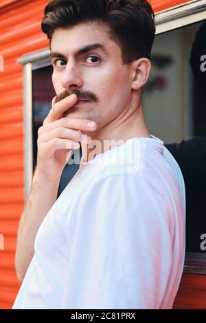 Portrait of young attractive mustache man in T-shirt dreamily looking in camera on street Stock Photo