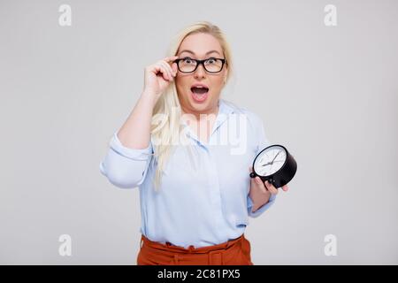 lateness concept - portrait of surprised beautiful plus size blonde woman holding alarm clock and screaming over gray background Stock Photo