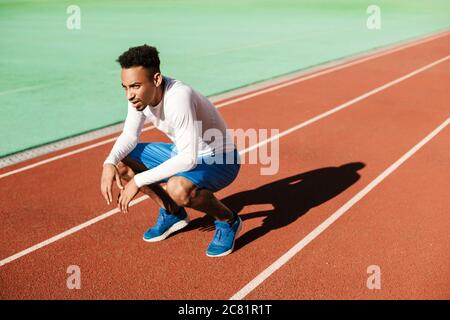 Young tired African American sportsman sit on one's haunches and resting after run at stadium Stock Photo